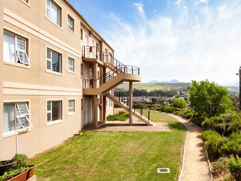 Rear view of the apartment building featuring an external staircase, landscaped garden, paved walkway, and scenic mountain views.