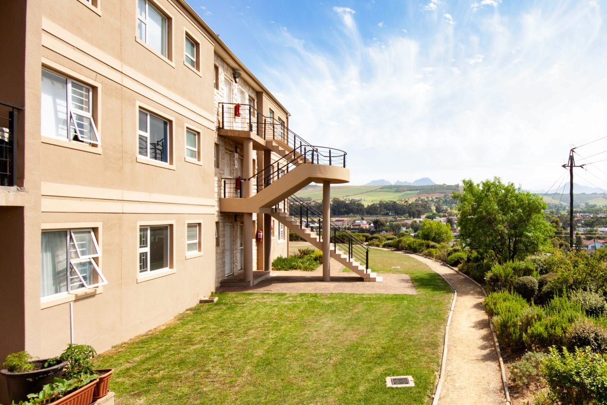 Rear view of the apartment building featuring an external staircase, landscaped garden, paved walkway, and scenic mountain views.