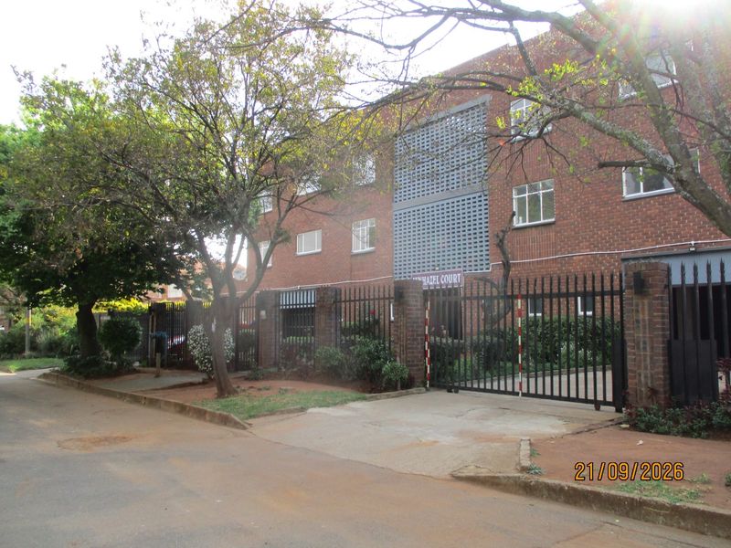 Front exterior view of a brick townhouse complex featuring a secure metal gate, concrete driveway, and decorative breeze block architectural detailing.