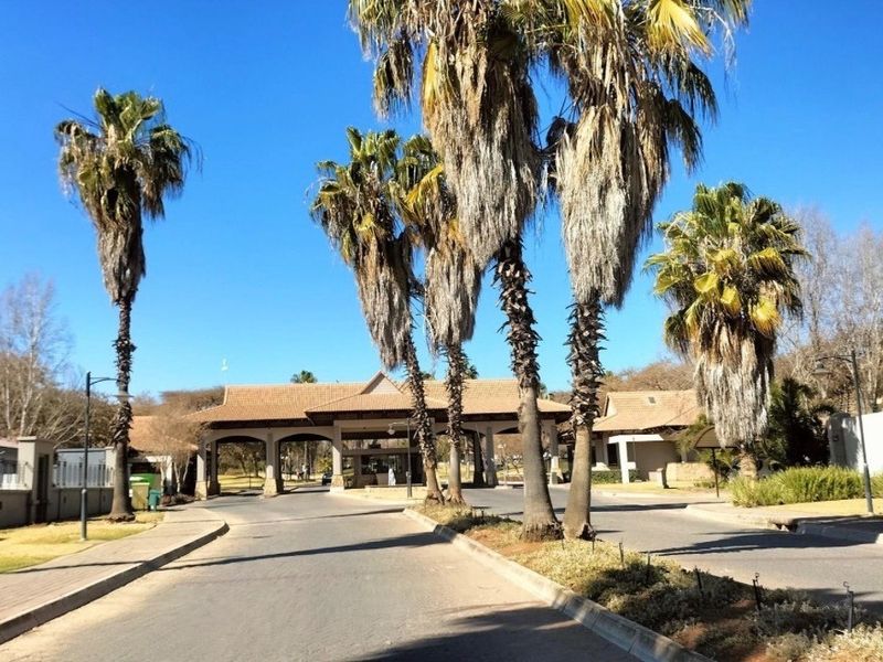 A view of the residential estate entrance featuring a paved road, mature palm trees, and a secure gatehouse structure.
