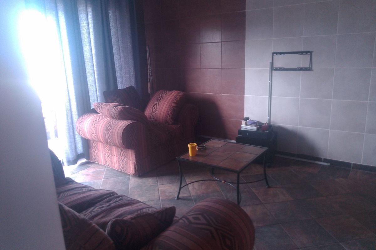 A living room area featuring tiled flooring and a tiled feature wall with a mounted television bracket, illuminated by natural light from a window.