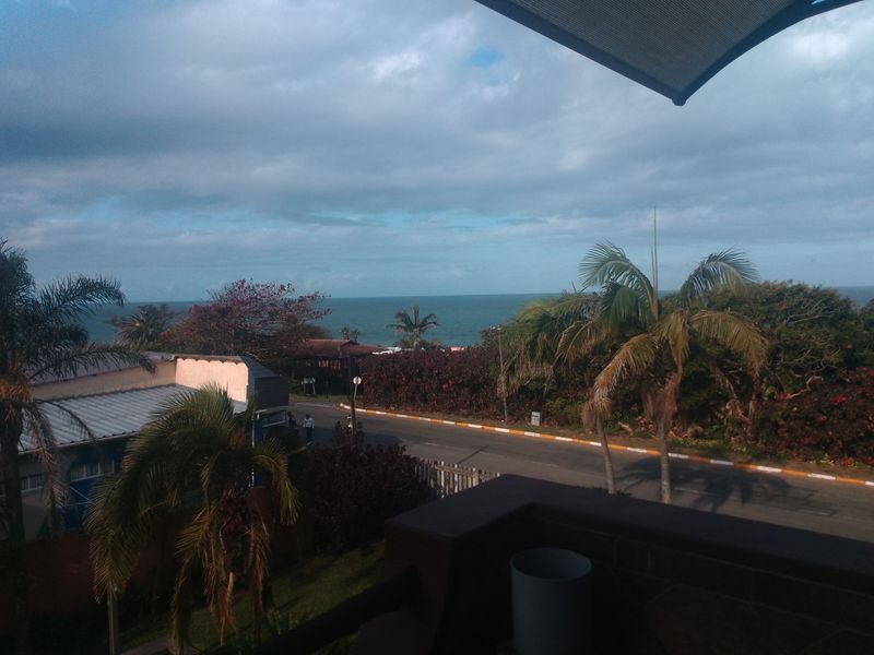 An elevated view from a balcony overlooking a coastal road, surrounding vegetation, and the ocean horizon.