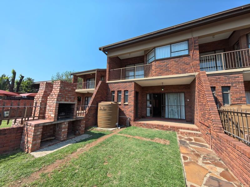 Rear view of a double-story brick house featuring a built-in brick braai, a water storage tank, a paved walkway, and an upper-level balcony.