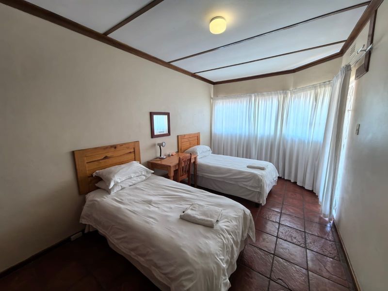 A bedroom featuring terracotta tile flooring and an exposed beam ceiling, offering a functional space with natural light through large windows.