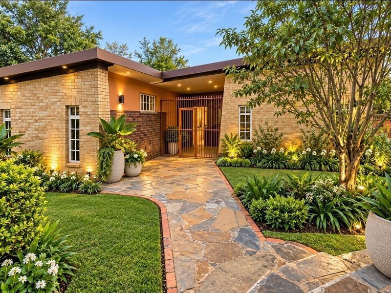 A beautifully landscaped front entrance featuring a stone paved walkway, brick facade, and secure metal gate with integrated exterior lighting.