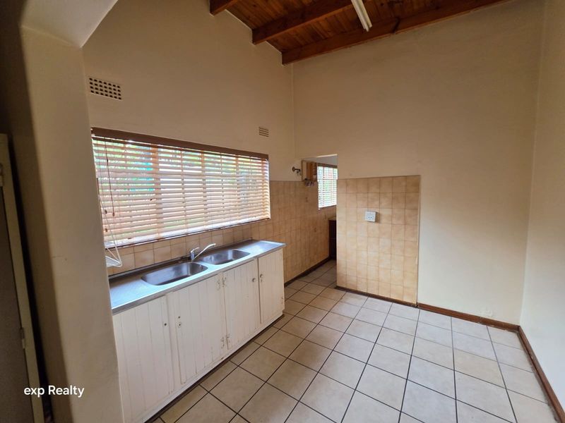 A functional kitchen space featuring tiled flooring, exposed timber ceiling beams, built-in base cabinetry with a double stainless steel sink, and tiled splashbacks.