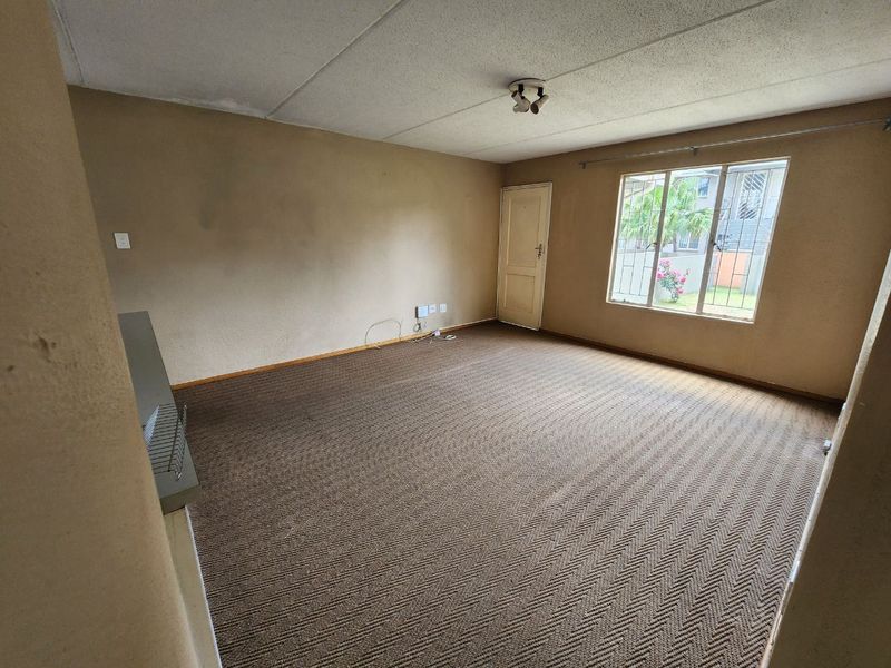 An empty living room featuring carpet flooring, ceiling tiles, and a window with security bars, presenting a blank canvas for renovation.
