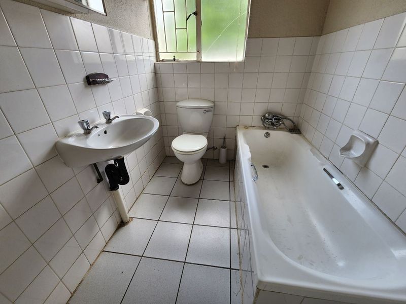 A dated bathroom featuring a pedestal basin, toilet, and bathtub with white ceramic wall and floor tiles, requiring cosmetic updates and maintenance.