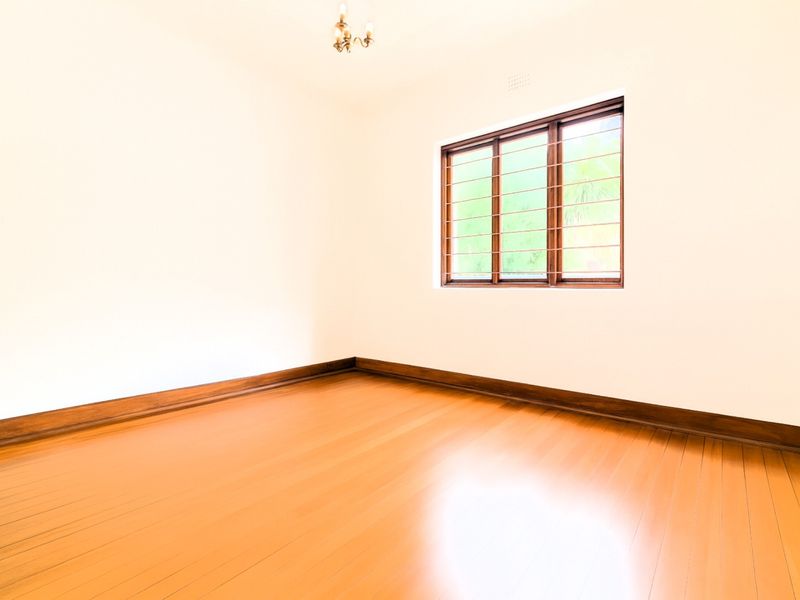 A bright, empty bedroom featuring polished timber flooring, timber-framed windows with security bars, and classic timber skirting boards.