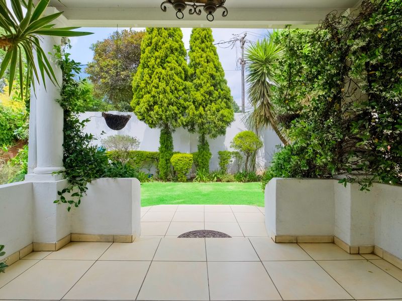 A well-maintained covered patio featuring tiled flooring and structural columns, overlooking a neat, landscaped rear garden with established trees and a boundary wall.