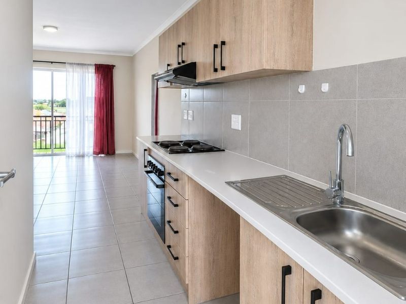 A modern galley-style kitchen featuring light wood-look cabinetry, stainless steel sink, built-in oven, and hob, with neutral tiled flooring throughout.
