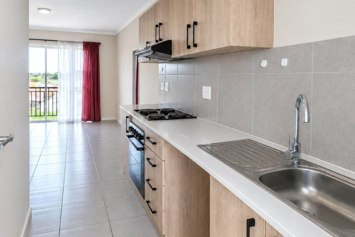 A modern galley-style kitchen featuring light wood-look cabinetry, stainless steel sink, built-in oven, and hob, with neutral tiled flooring throughout.