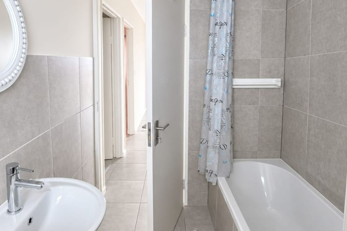 A clean, neutral-toned bathroom featuring a pedestal basin, a standard bathtub with a shower curtain rail, and modern ceramic tiling throughout.
