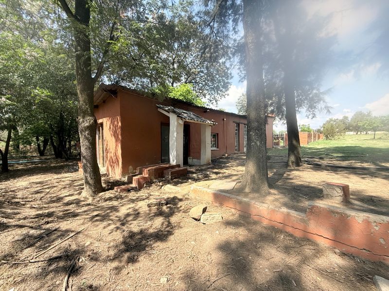 Rear view of a rustic farm building featuring brick construction, a tiled roof, and a low retaining wall set within a wooded garden area.