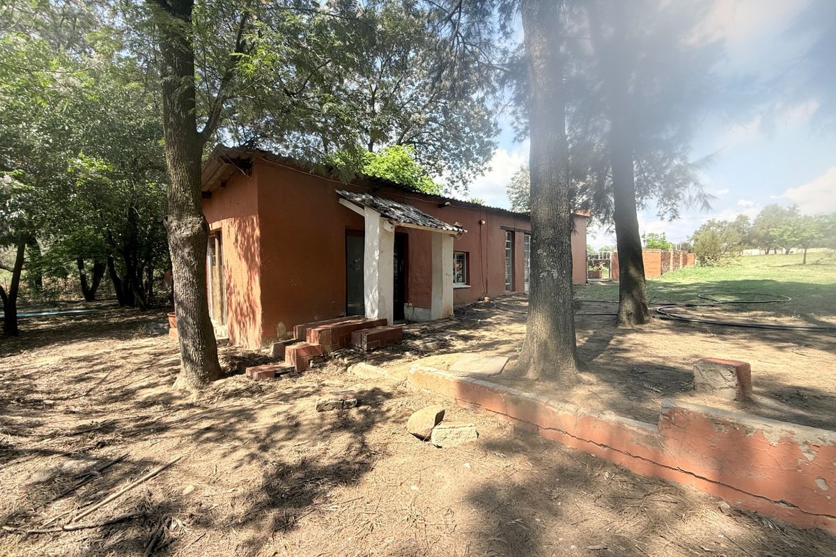 Rear view of a rustic farm building featuring brick construction, a tiled roof, and a low retaining wall set within a wooded garden area.