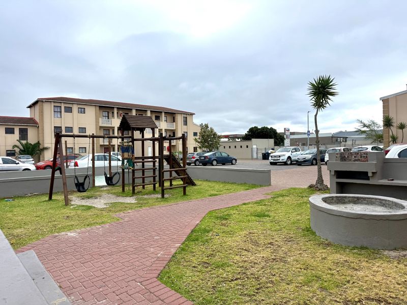 Communal outdoor area featuring a paved walkway, built-in braai facility, and playground equipment set within a residential apartment complex.