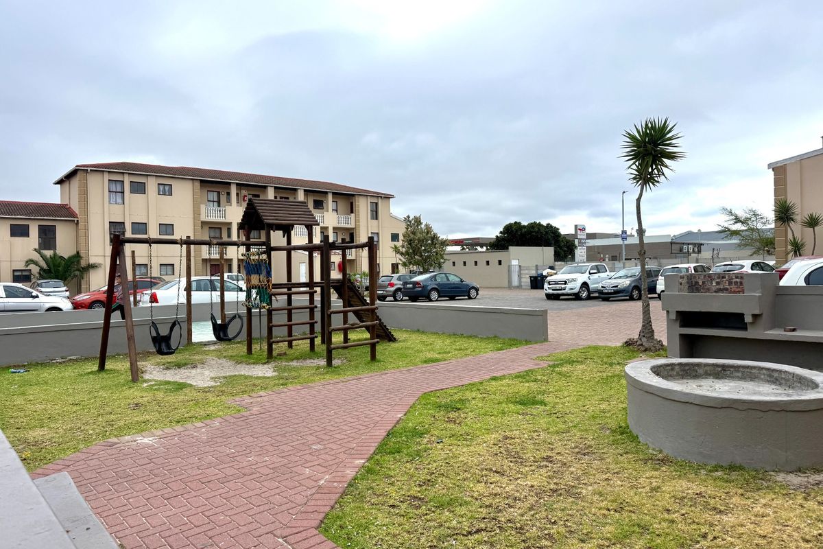 Communal outdoor area featuring a paved walkway, built-in braai facility, and playground equipment set within a residential apartment complex.