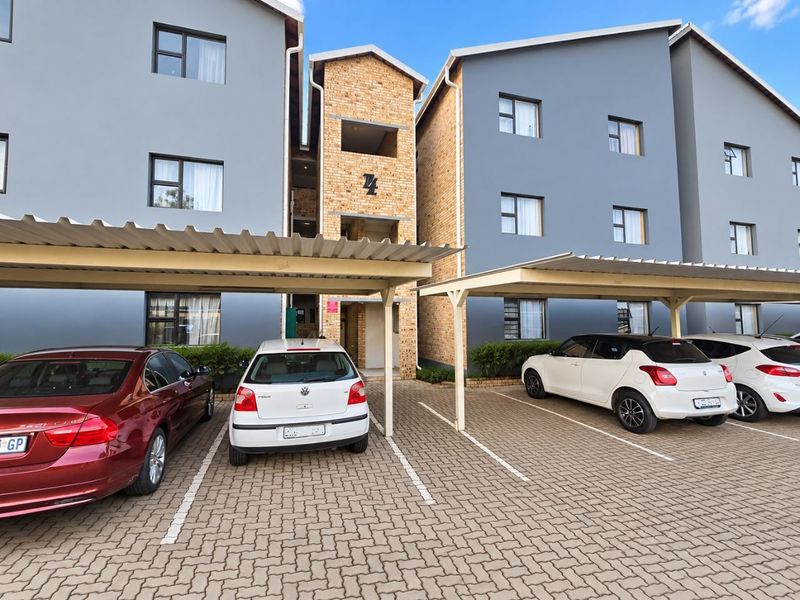 Front exterior view of a multi-story townhouse complex featuring covered parking bays, paved driveway, and a combination of brick and painted plaster finishes.