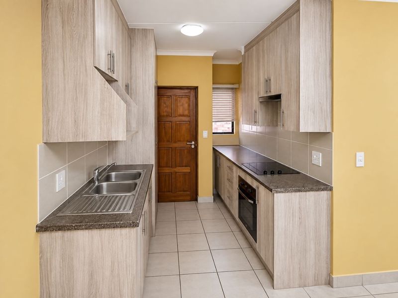 A clean, galley-style kitchen featuring light wood-grain cabinetry, dark laminate countertops, integrated oven and hob, and neutral tiled flooring.