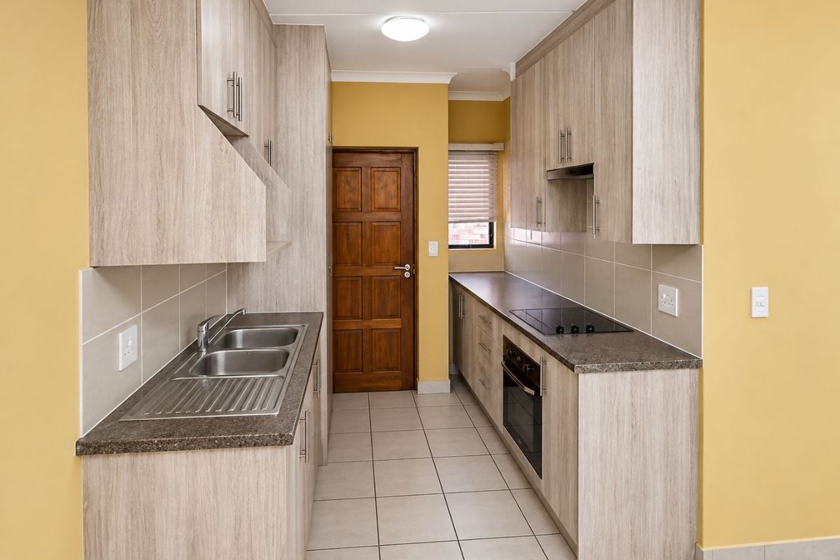 A clean, galley-style kitchen featuring light wood-grain cabinetry, dark laminate countertops, integrated oven and hob, and neutral tiled flooring.