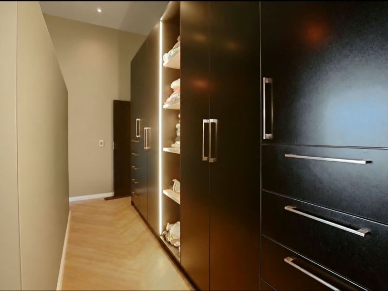 A modern walk-in wardrobe featuring dark custom cabinetry with integrated LED lighting and high-quality herringbone timber flooring.