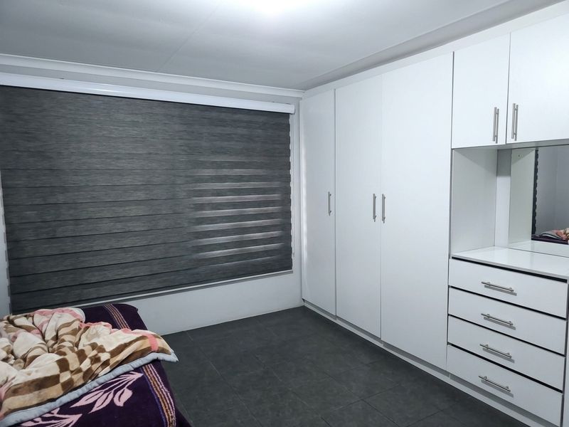A well-maintained bedroom featuring modern white built-in cupboards with an integrated vanity, dark tiled flooring, and contemporary grey roller blinds.
