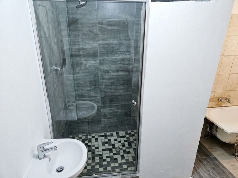 A functional bathroom featuring a glass-enclosed shower with grey tiling, a pedestal basin, and a separate bathtub with visible wear.