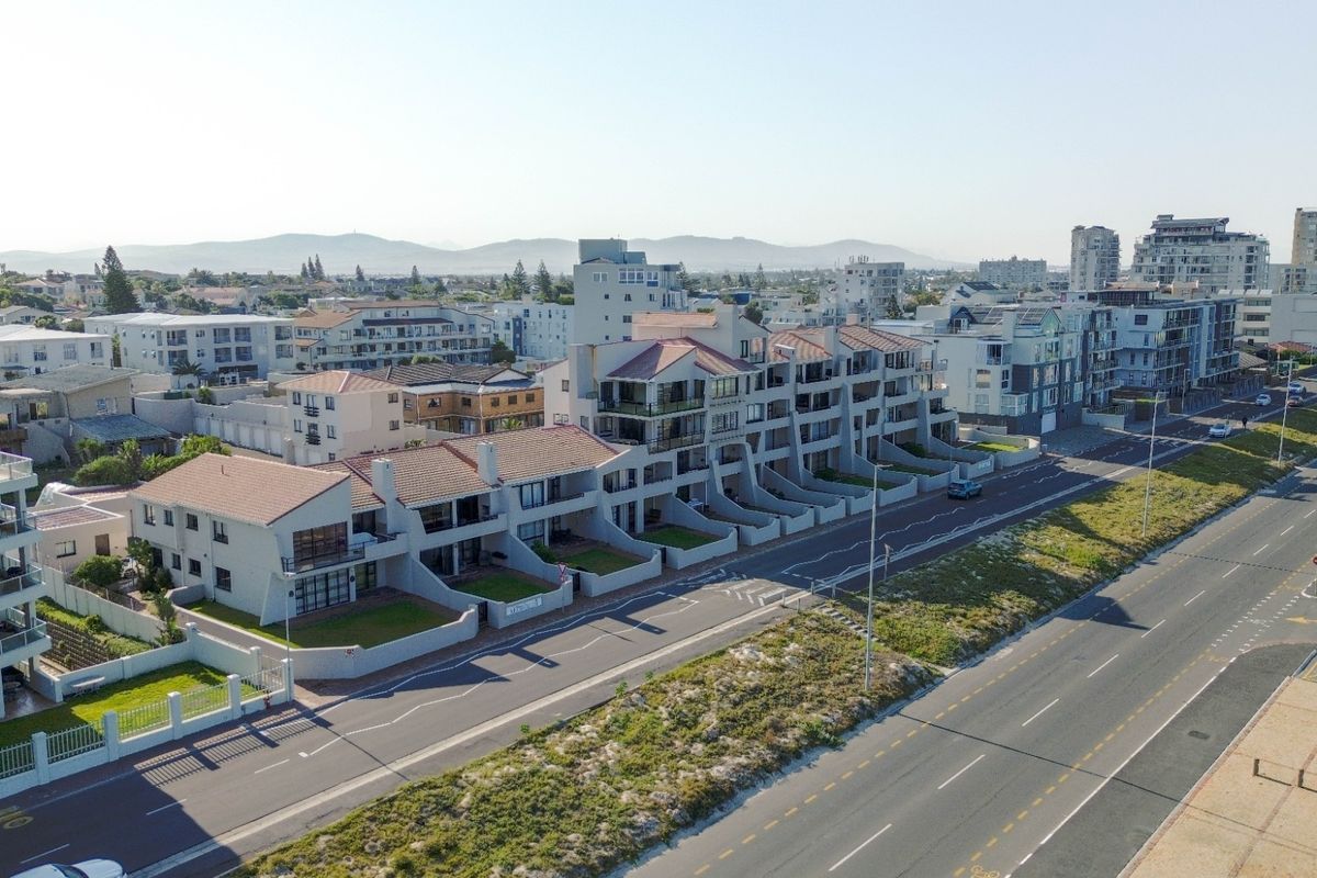 Aerial view of a coastal apartment complex featuring multi-level units with tiled roofs, balconies, and private garden spaces along a main road.
