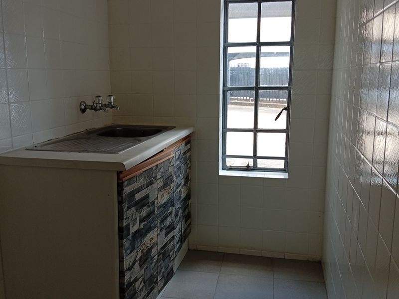 A compact scullery area featuring tiled walls, a stainless steel sink, and a window with security bars, offering a functional utility space.