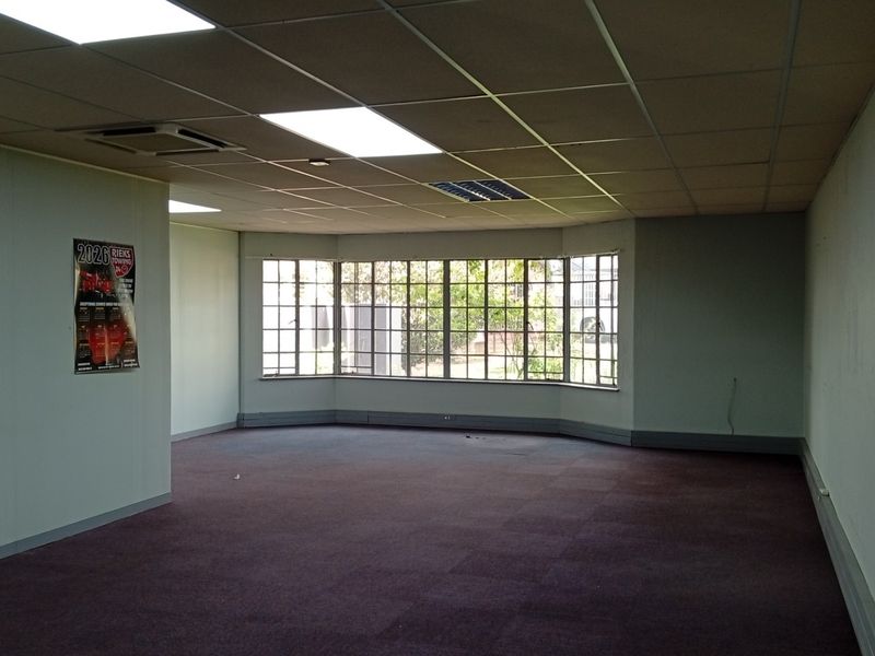 An empty, open-plan office space featuring a large bay window, suspended ceiling with recessed lighting, and carpeted flooring.