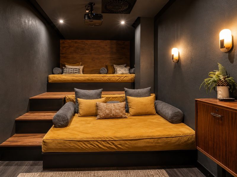 A modern, professionally designed cinema room featuring tiered seating platforms, integrated projector, and ambient wall sconce lighting.