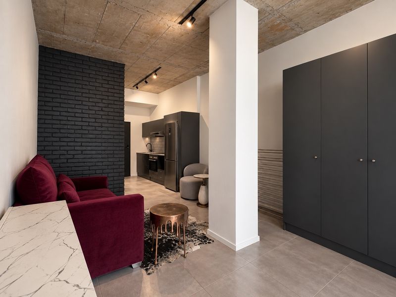 Modern open-plan living area featuring an exposed concrete ceiling, industrial-style track lighting, a sleek kitchen with built-in appliances, and a large integrated wardrobe.