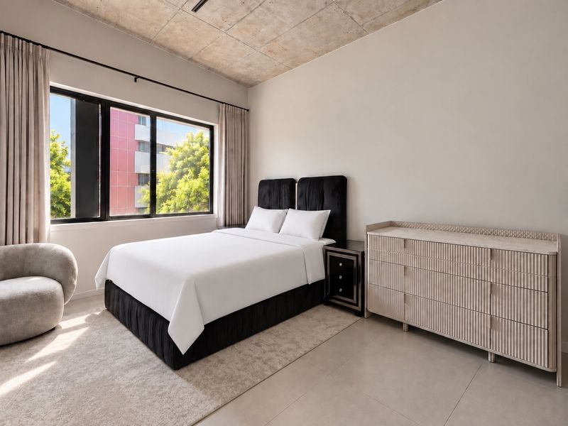 A modern bedroom featuring an exposed concrete ceiling, large window with natural light, and neutral tiled flooring in a well-maintained apartment setting.