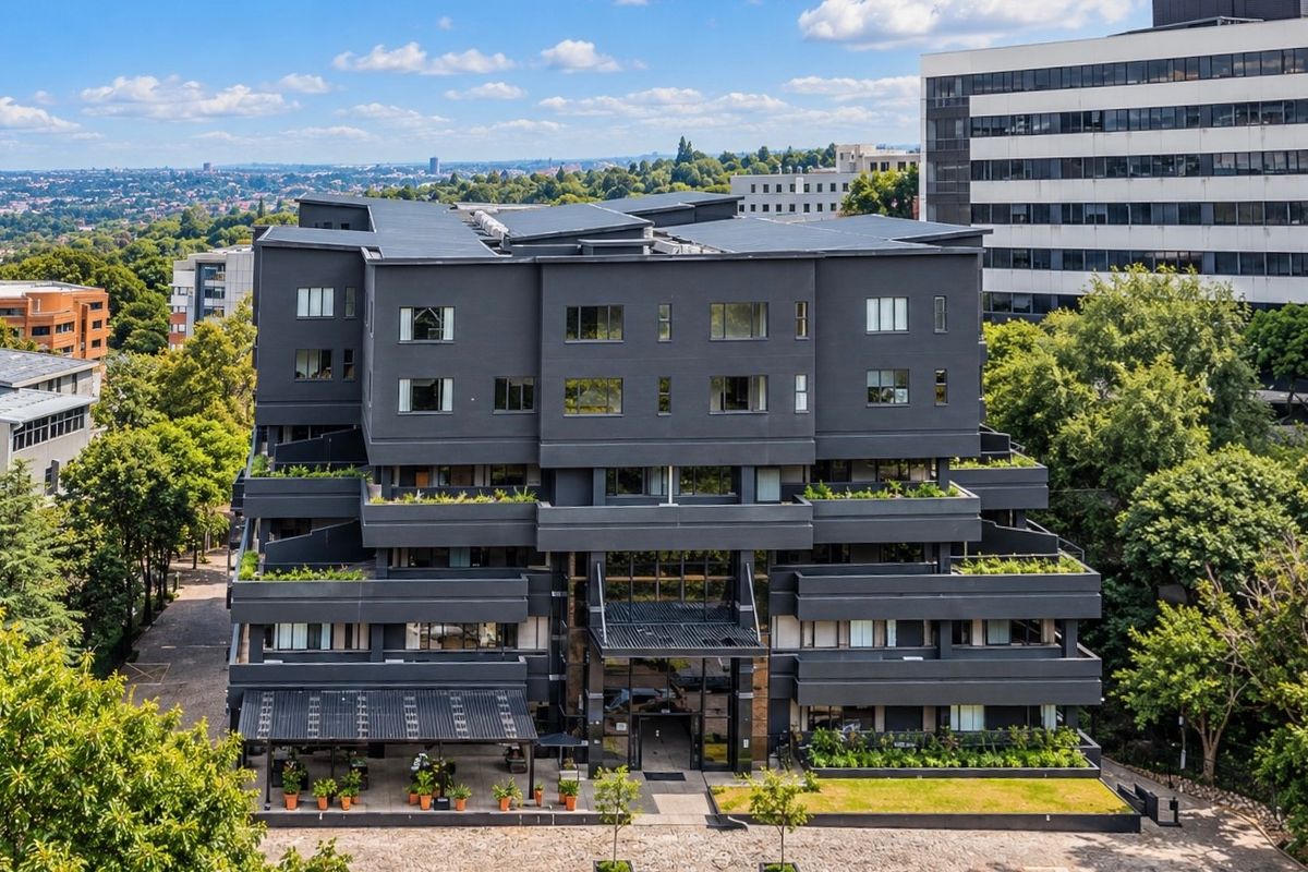 Aerial view of a modern multi-story apartment building featuring dark exterior cladding, tiered balconies with greenery, and a paved ground-level entrance area.