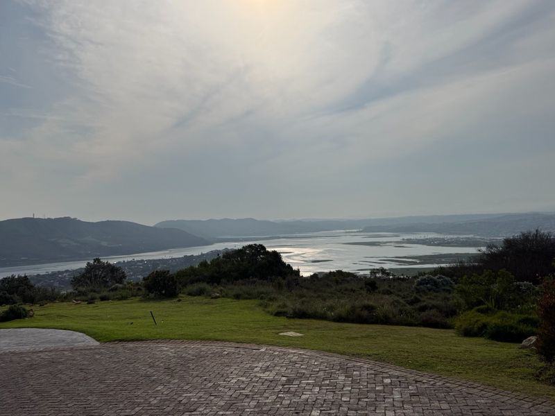A scenic elevated view overlooking a river estuary and surrounding hills, featuring a paved driveway and well-maintained lawn in the foreground.