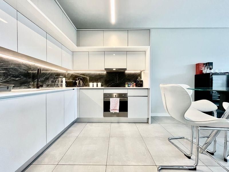 Modern kitchen featuring sleek white cabinetry, dark stone splashback, integrated oven, and high-quality tiled flooring with ambient under-cabinet lighting.