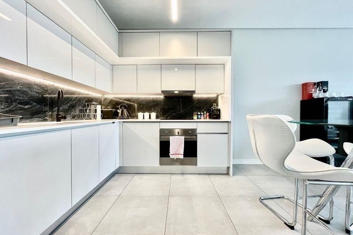 Modern kitchen featuring sleek white cabinetry, dark stone splashback, integrated oven, and high-quality tiled flooring with ambient under-cabinet lighting.