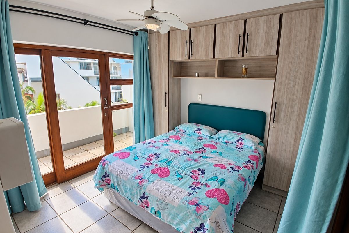 A well-lit bedroom featuring built-in wooden cupboards, a ceiling fan, tiled flooring, and direct access to a private balcony through sliding glass doors.