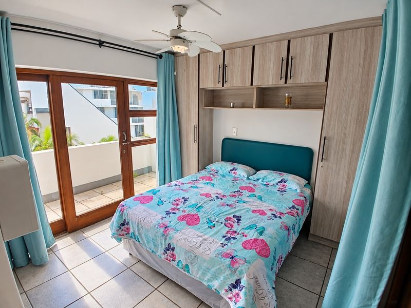 A well-lit bedroom featuring built-in wooden cupboards, a ceiling fan, tiled flooring, and direct access to a private balcony through sliding glass doors.