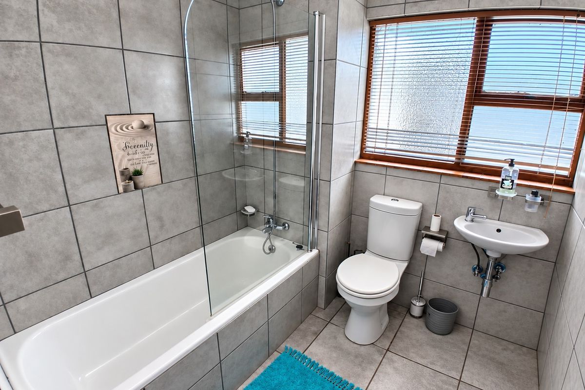 A modern bathroom featuring a bathtub with a glass shower screen, a wall-mounted basin, and a toilet, finished with neutral grey floor-to-ceiling tiles.