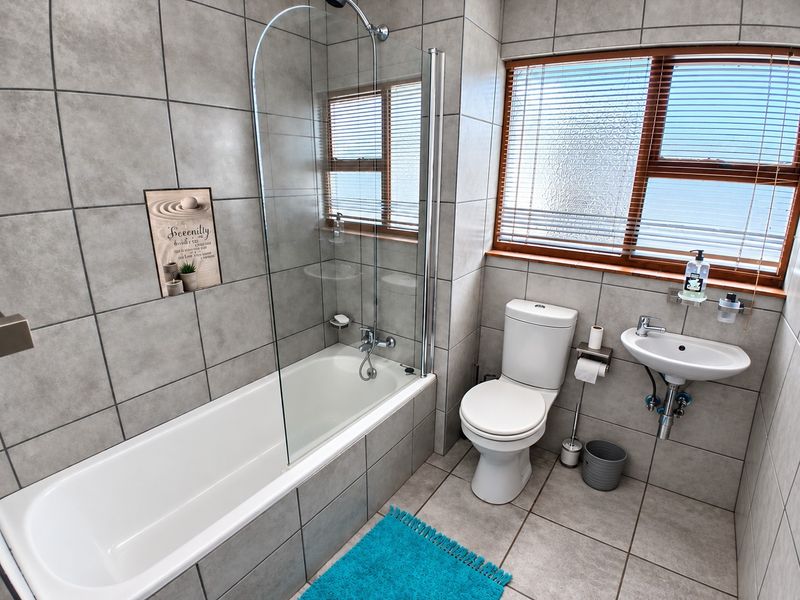 A modern bathroom featuring a bathtub with a glass shower screen, a wall-mounted basin, and a toilet, finished with neutral grey floor-to-ceiling tiles.