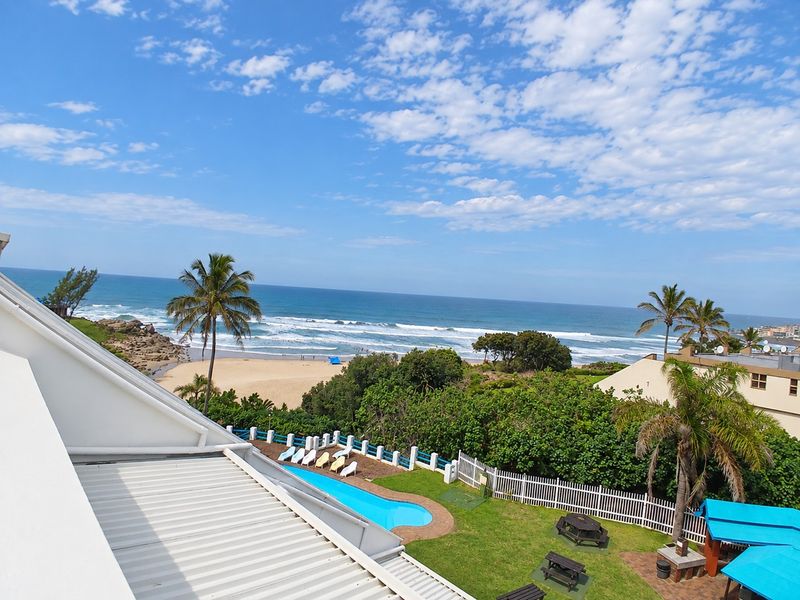 Elevated coastal view overlooking the swimming pool, garden area, and the beach with ocean waves in the background.