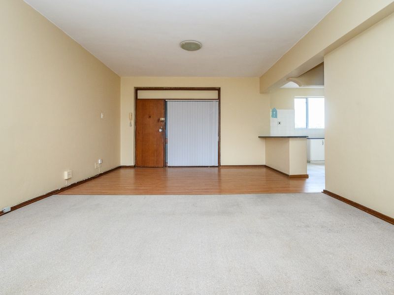 An empty open-plan living area featuring a mix of carpet and laminate flooring, with a breakfast bar connecting to the kitchen space.