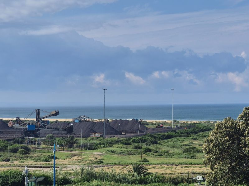 An elevated view overlooking a coastal landscape featuring industrial infrastructure, coal stockpiles, and the ocean in the distance.