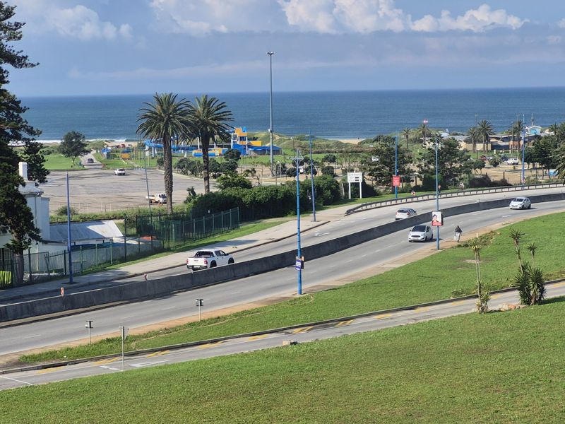 An elevated view overlooking a coastal road, green spaces, and the ocean in the distance.