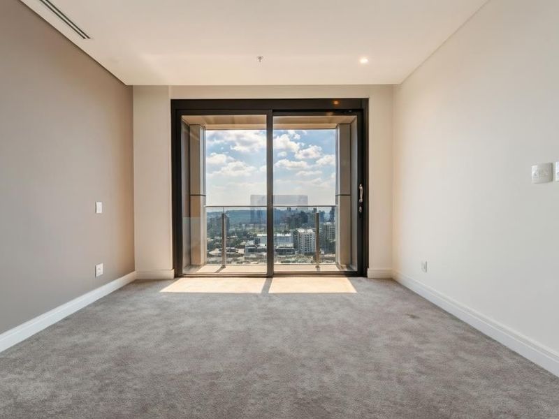A bright, empty bedroom featuring plush carpeting, recessed lighting, and a large sliding glass door leading to a balcony with city views.