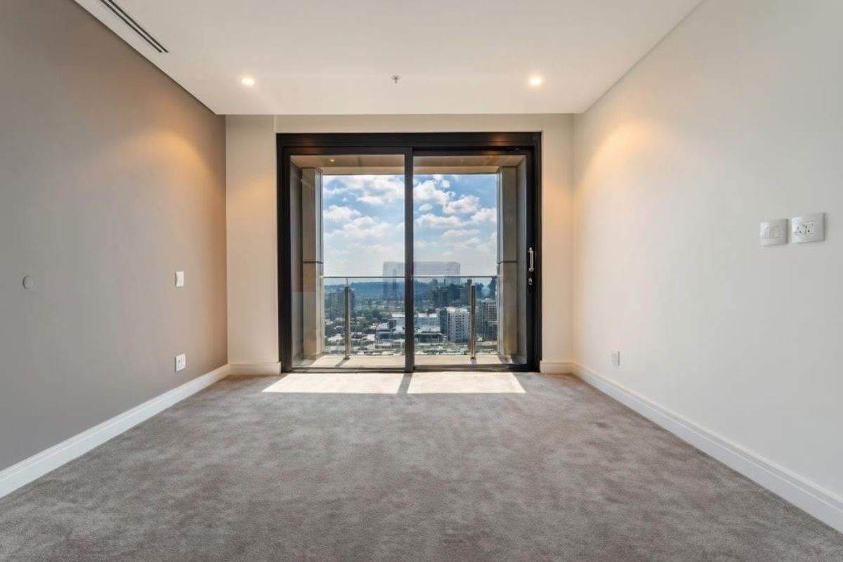 A bright, empty bedroom featuring plush carpeting, recessed lighting, and a large sliding glass door that opens to a balcony with city views.