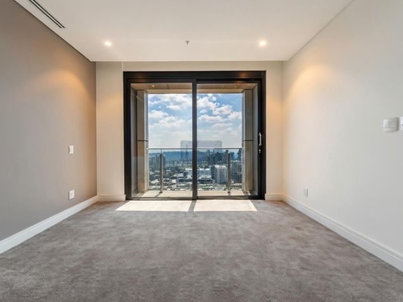 A bright, empty bedroom featuring plush carpeting, recessed lighting, and a large sliding glass door that opens to a balcony with city views.