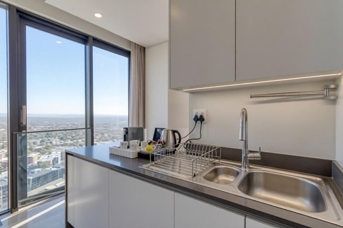 Modern kitchen area featuring sleek grey cabinetry, dark stone countertops, a stainless steel double sink, and floor-to-ceiling windows with expansive city views.