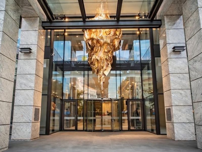 Grand entrance of the apartment building featuring marble-clad pillars, a glass canopy, and a striking sculptural light fixture.